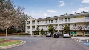 A multi-story hotel with a parking lot, a sign indicating a bank branch, and a clear sky. - Suburban Studios Winston Salem-University Parkway (Winston-Salem)