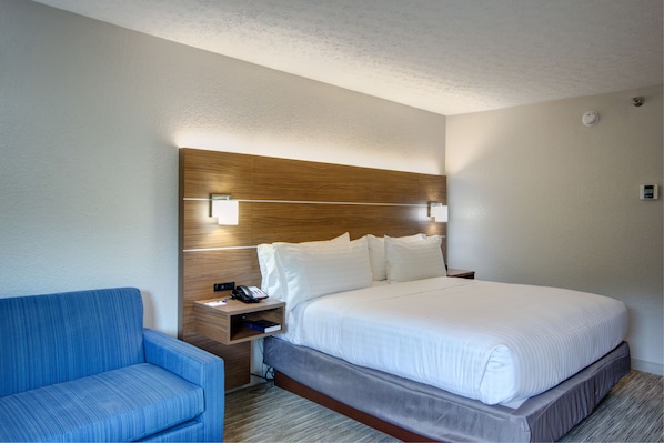 Une chambre d’hôtel avec un grand lit, une chaise bleue, une tête de lit en bois, des lampes de chevet et une table de chevet avec un téléphone et un livre.