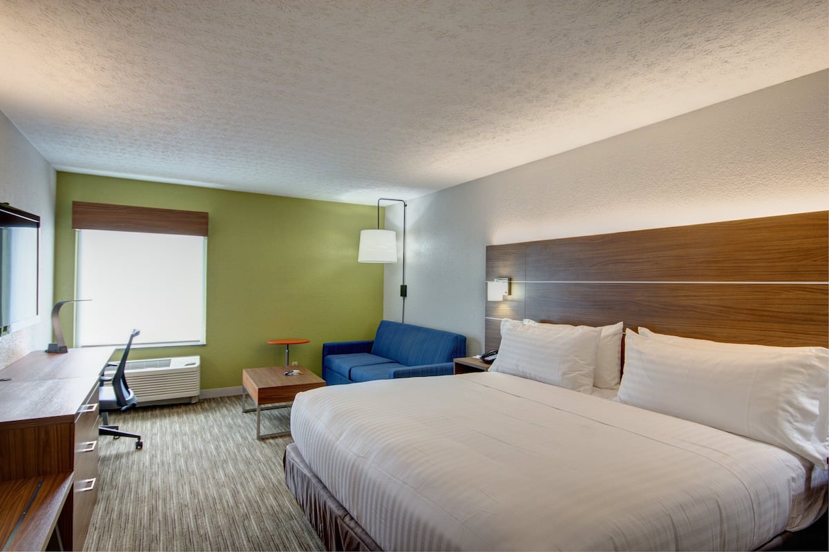 A hotel room with a large bed, a desk with a chair, a blue sofa, and a bedside table with a lamp.