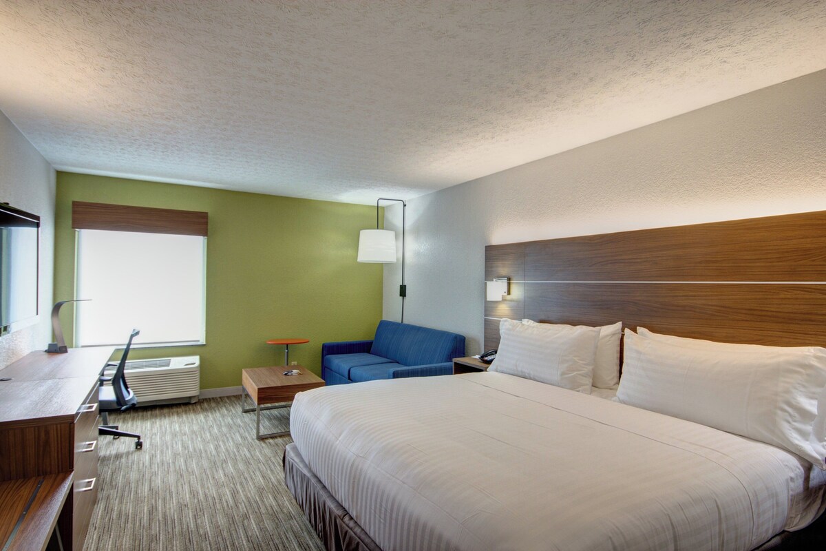 A hotel room with a bed, a desk, a chair, a lamp, and a window.