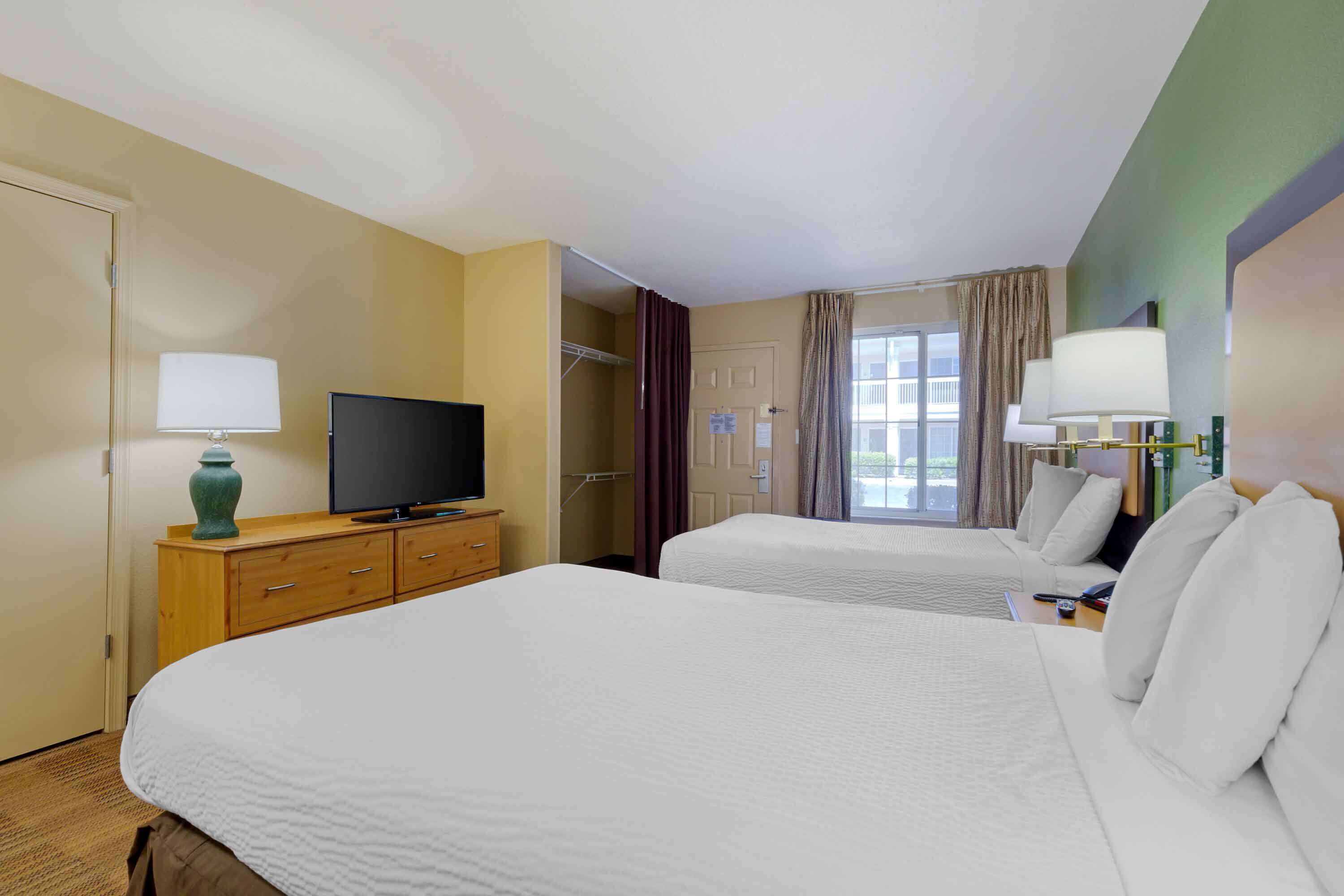 Habitación de hotel con dos camas, televisor, escritorio con lámpara y ventana con cortinas.