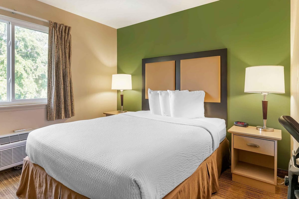 Ein Hotelzimmer mit einem großen Bett, zwei Nachttischen mit Lampen, einem Fenster mit Vorhängen und einer Wandheizung.