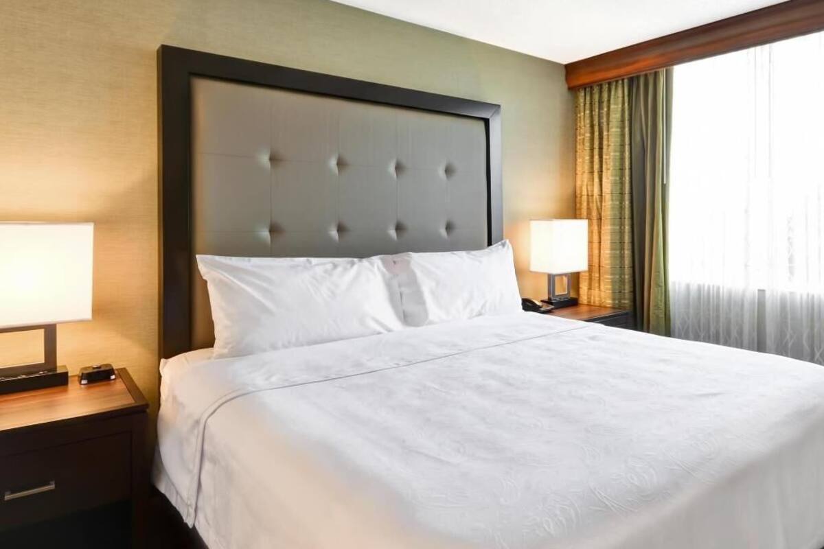A hotel room with a large bed, two bedside tables, a headboard, and a window with curtains.