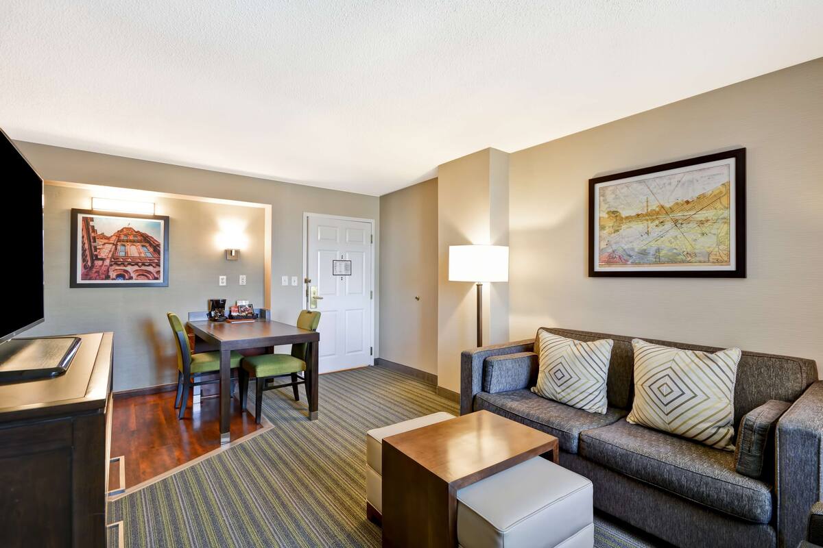 A hotel room with a dining area, a sofa set, a flat-screen TV, and two wall-mounted lamps.