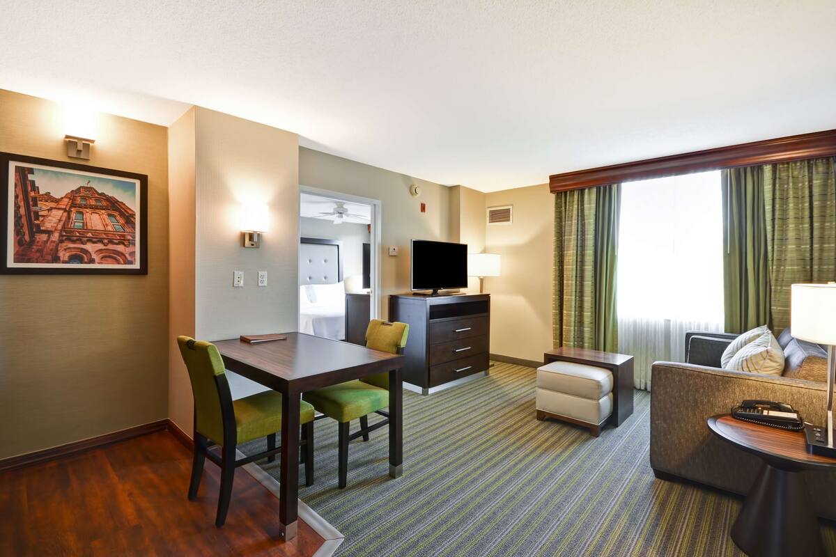 A hotel room with a dining table, chairs, a sofa, a TV, and a window with curtains.