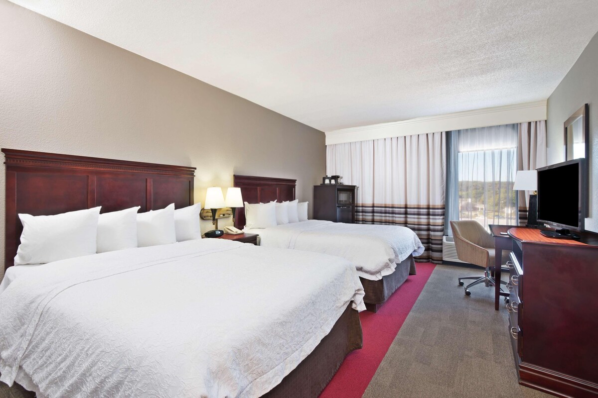 A hotel room with two beds, a desk, a television, and a window with curtains.