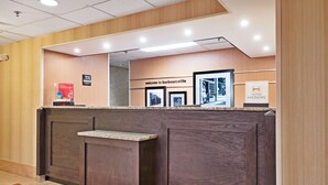 A reception desk with a dark wood finish, a welcome sign, framed pictures, and a Hilton Honors logo. - Hampton Inn Huntington/Barboursville (Barboursville)