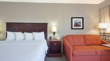 A hotel room with a large bed, a wooden nightstand with a lamp, a red sofa, and a framed picture on the wall.