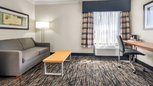Ein Hotelzimmer mit einer Couch, einem kleinen Tisch, einem Schreibtisch mit Stuhl, einer Lampe und einem Fenster mit Vorhängen.