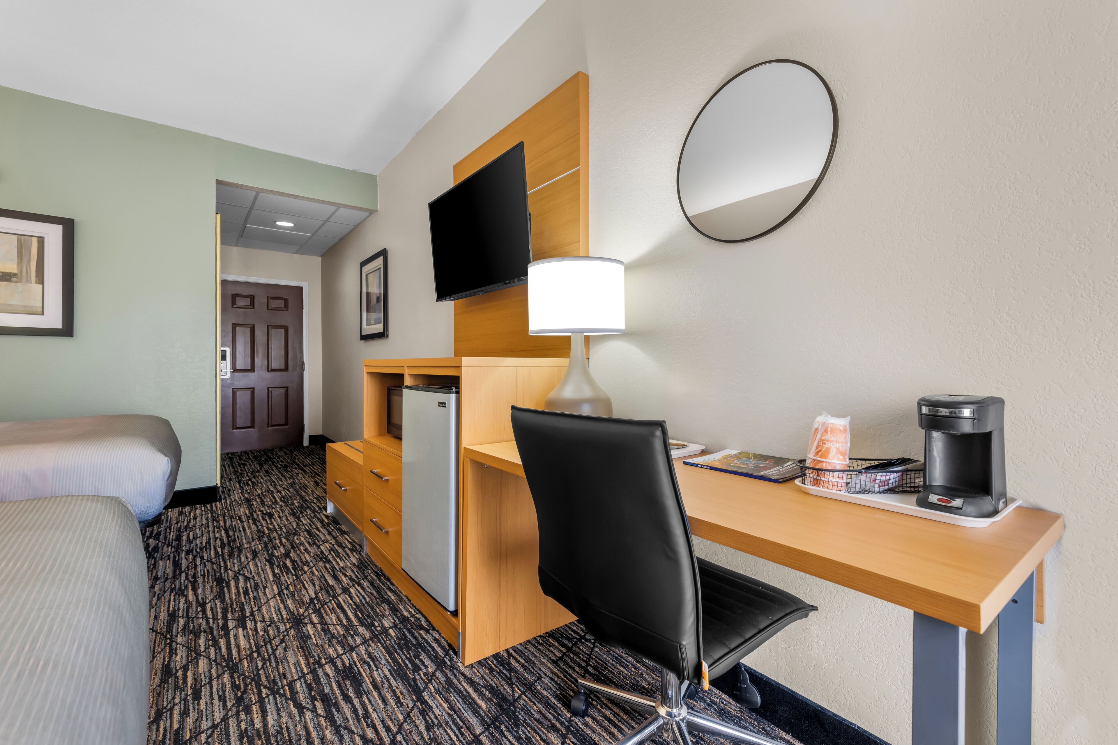 A hotel room with a bed, desk, chair, desk lamp, coffee maker, refrigerator, and a TV mounted on the wall.