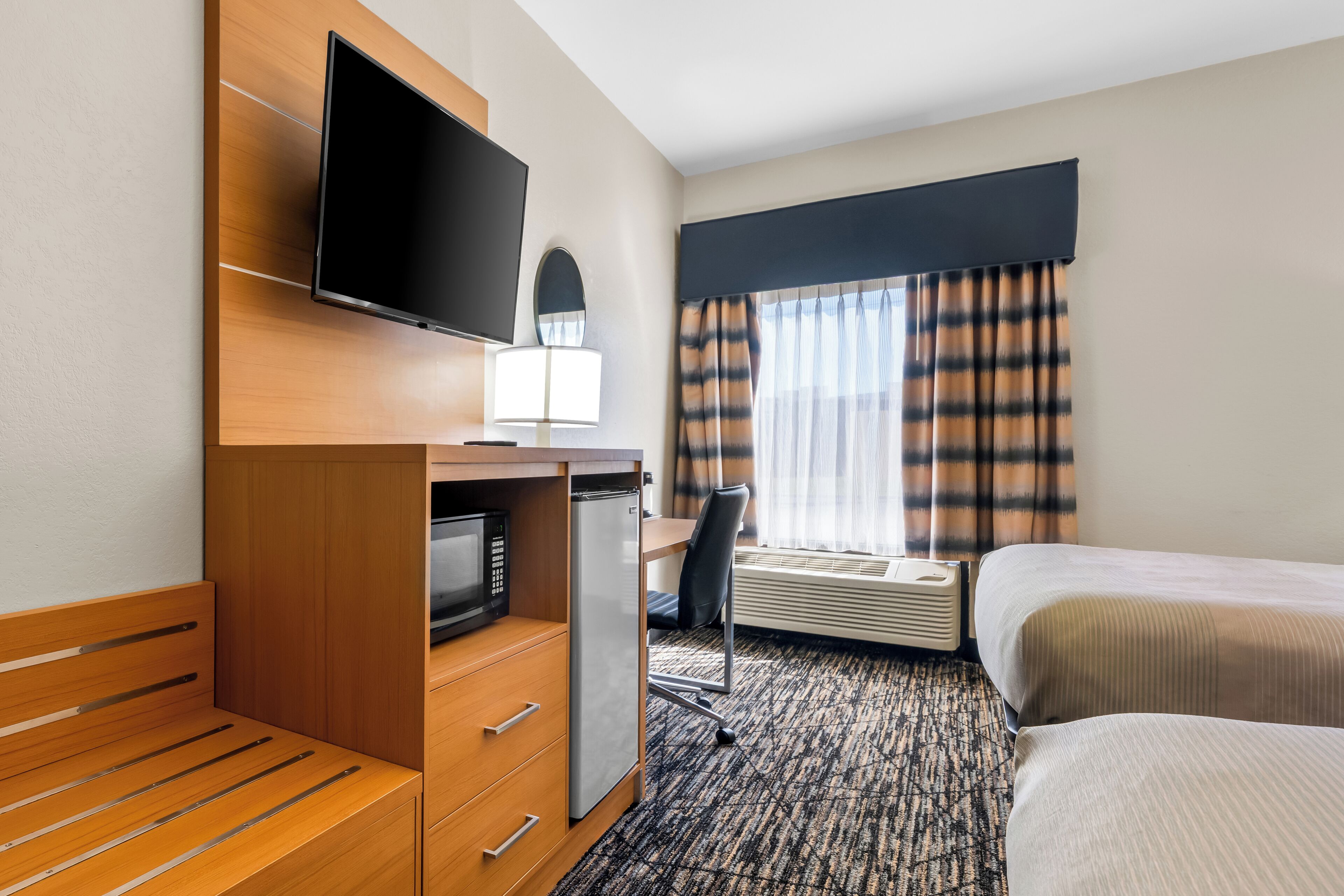 A hotel room with a bed, a desk with a television, a microwave, and a window with curtains.