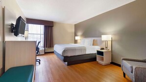 A hotel room with a bed, a desk with a TV, a chair, a bedside table with a lamp, and a sofa.