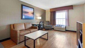 A hotel room with a sofa, a small table, a chair, a desk with a lamp, an air conditioning unit, and a large window with curtains.