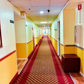 A long, narrow hallway with yellow walls and a red carpet. There are doors on both sides, and a sign indicating "Rooms 11-24" on the left.