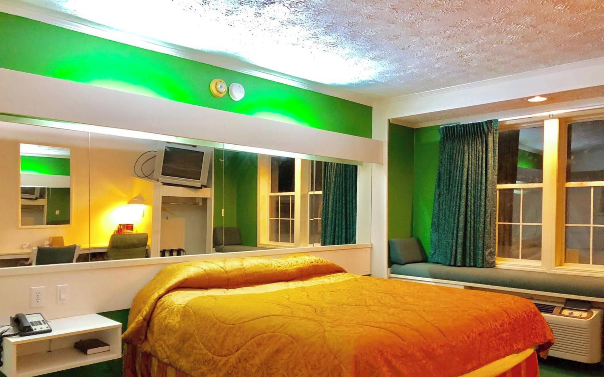 Quarto de hotel com cama, escrivaninha com telefone, televisão e janela com cortinas.