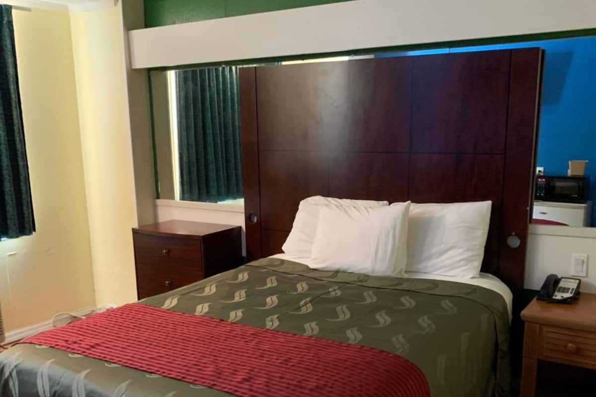 Habitación de hotel con una cama, una mesita de noche de madera, un escritorio, un teléfono y una ventana con cortinas.