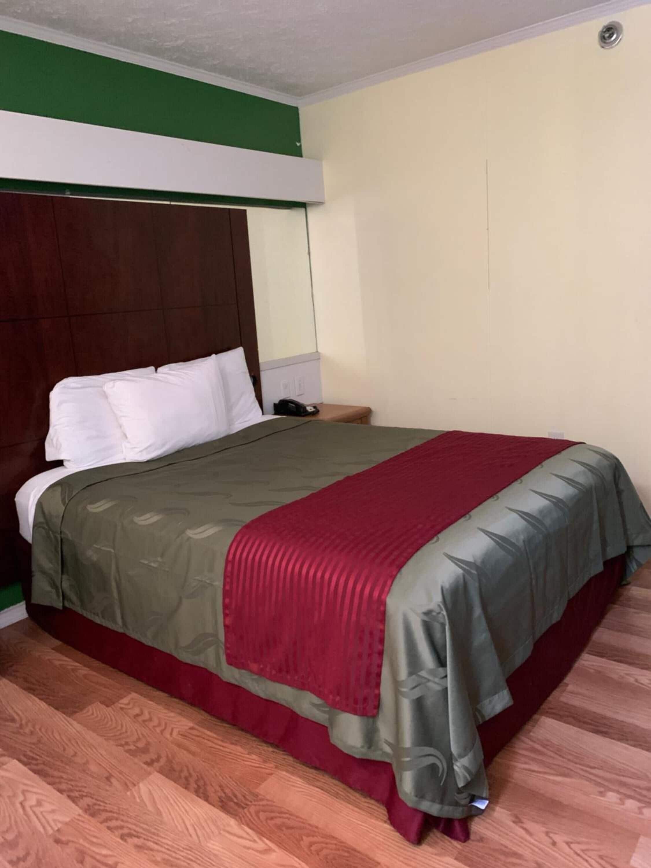 Habitación de hotel con cabecera de madera, cama con ropa de cama blanca y verde, una manta roja y suelo de madera.