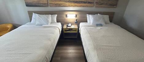 Habitación de hotel con dos camas, una mesita de noche con teléfono, una lámpara y un cuadro de un atardecer sobre el agua.