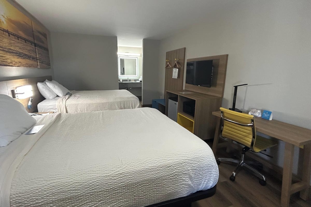 A hotel room with two beds, a desk, a chair, a TV, and a bathroom in the background.