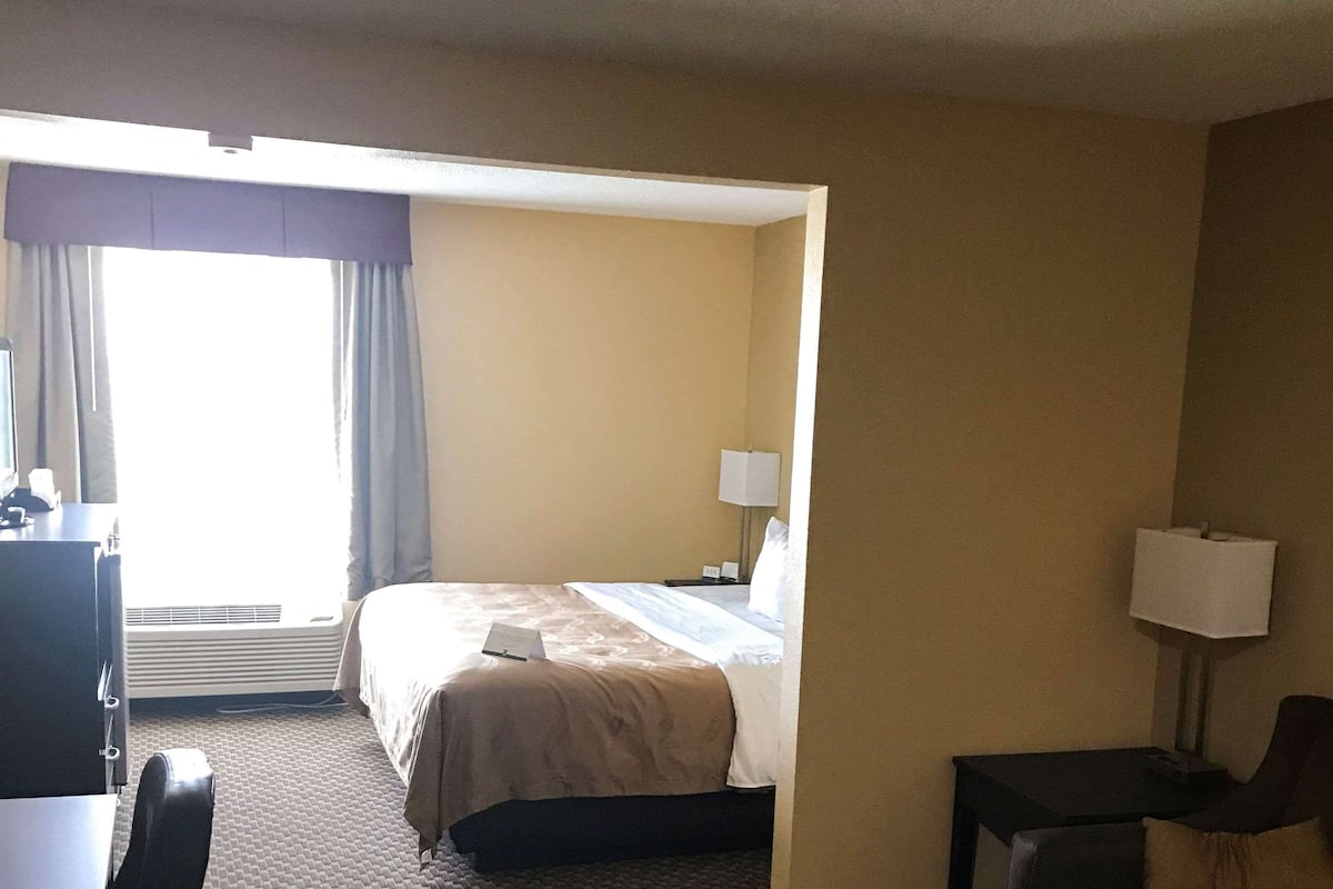 Ein Hotelzimmer mit Bett, Schreibtisch, Stuhl, Lampe und einem fensterbelaubten Fenster.