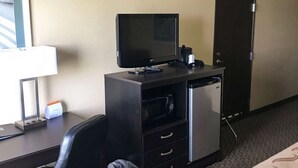 A hotel room with a desk, a flat-screen TV, a microwave, a mini-fridge, a chair, and a trash bin.