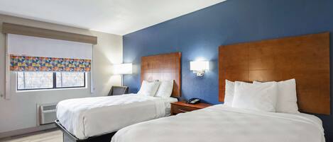 Quarto de hotel com duas camas, uma janela com persianas, uma luminária na parede e uma mesa de cabeceira com telefone.