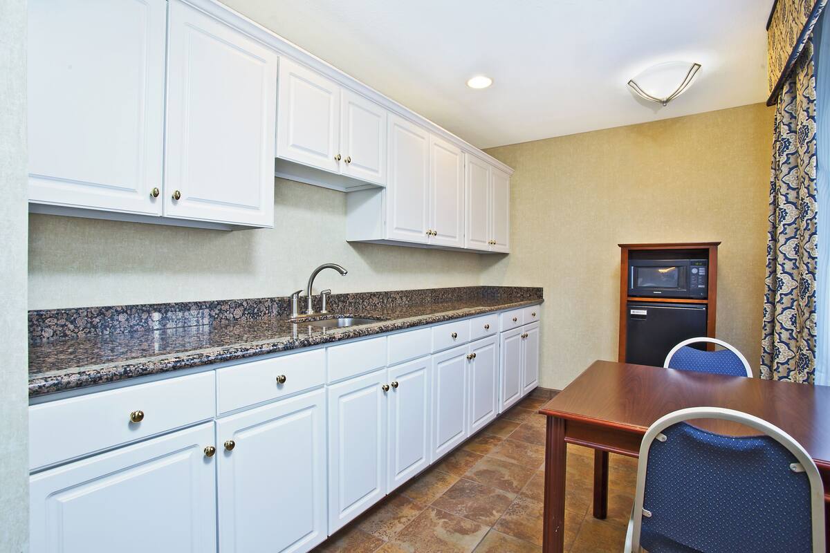 A compact kitchen with white cabinets, a granite countertop, and a small dining area with a wooden table and blue chairs.