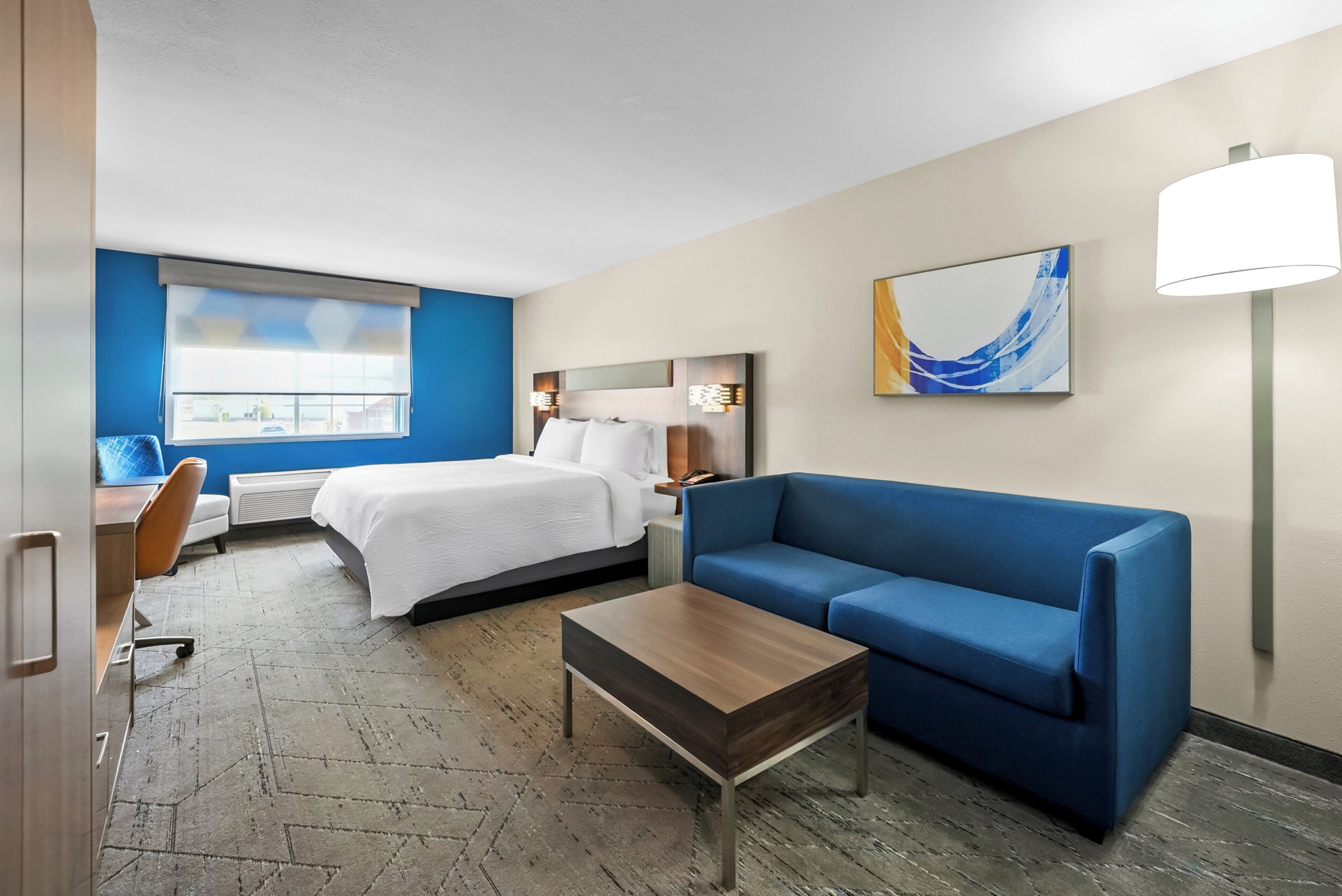 A hotel room with a bed, a blue sofa, a wooden coffee table, a desk with a chair, and a window with blinds.
