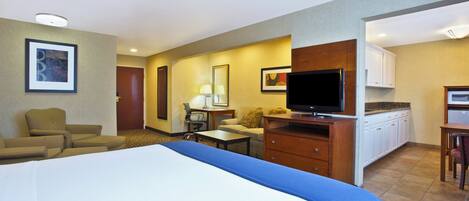 Quarto de hotel com cama, escrivaninha, televisão, cadeira, sofá e cozinha compacta.