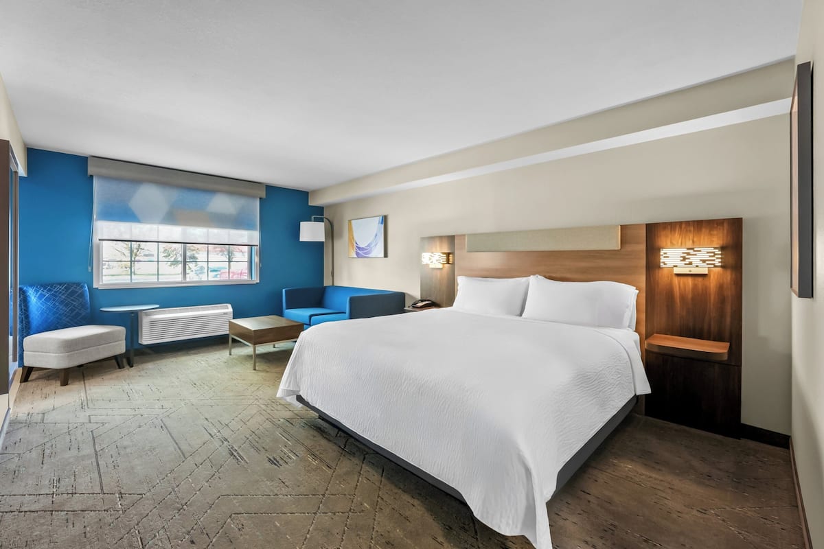 A hotel room with a large bed, a seating area with a chair and a table, a window with blinds, and a wall-mounted light fixture.