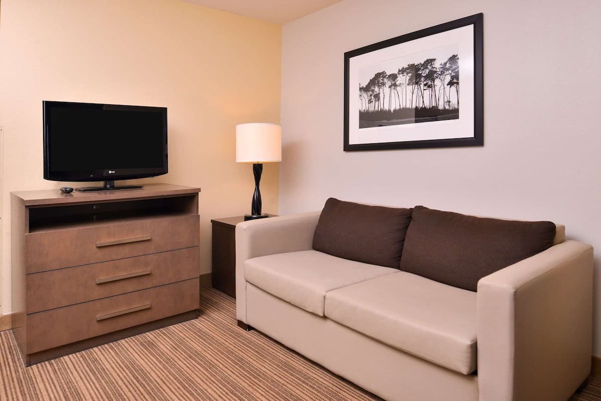 Ein Hotelzimmer mit Flachbildfernseher auf einer Holzkommode, einem beigen Sofa mit braunen Kissen, einem Beistelltisch mit Lampe und einem gerahmten Bild an der Wand.