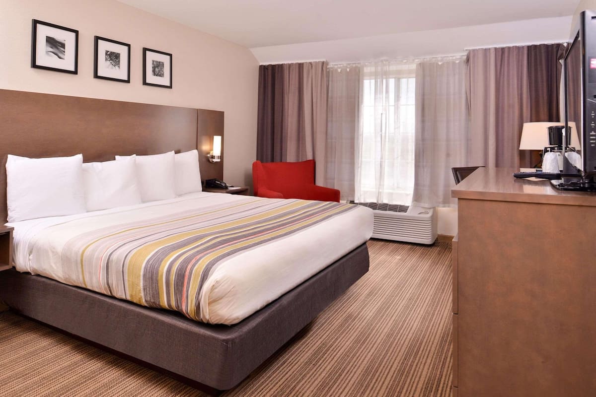 Ein Hotelzimmer mit einem großen Bett, einem Schreibtisch, einem Sessel und einem Fernseher. Vorhänge und ein Fenster sind vorhanden.