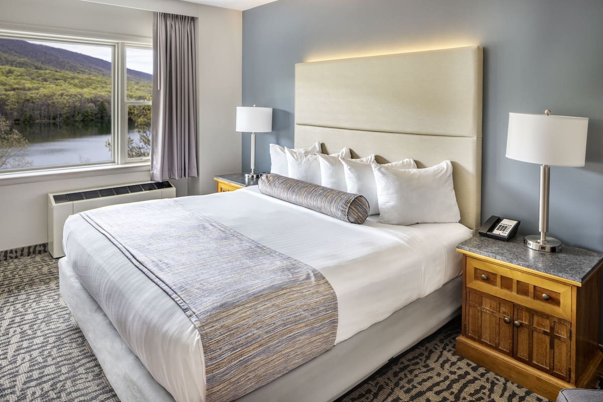 A hotel room with a large bed, two bedside tables with lamps, a view of a lake, and a wooden nightstand.