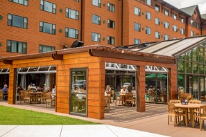 An outdoor dining area with wooden tables and chairs, a glass enclosure, and a building with multiple windows in the background. - Rocky Gap Casino & Resort (Cumberland)