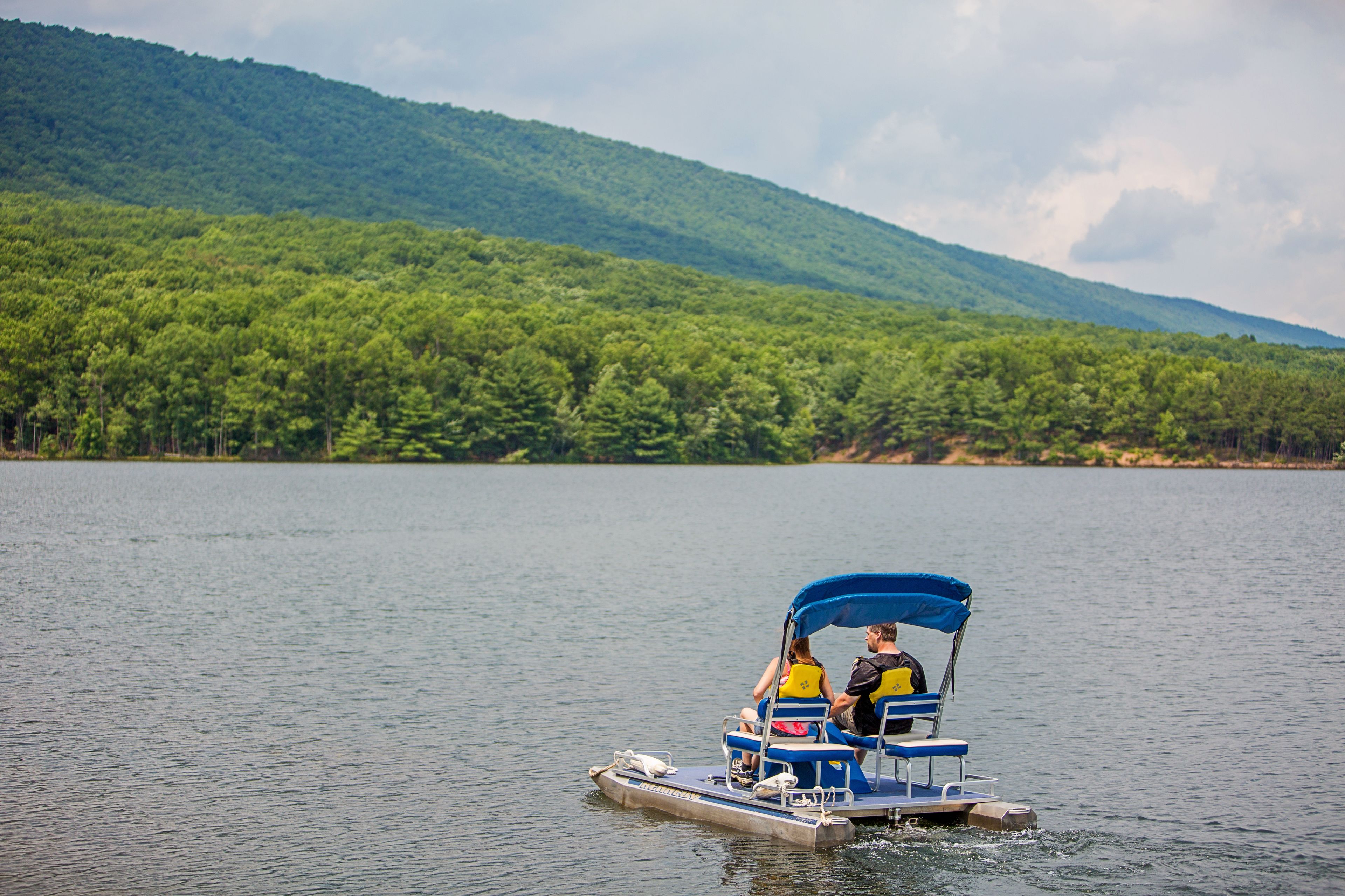 A small boat with two people on it, moving on a calm lake with a forested hill in the background.
