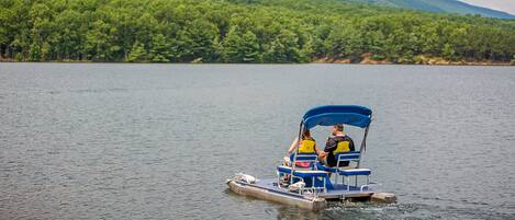 Una pequeña embarcación con dos personas navegando en un lago tranquilo, con una colina boscosa al fondo.