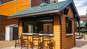 Un coin bar en bois, avec deux tabourets et un comptoir, situé à côté d’un bâtiment doté d’un toit végétalisé et d’une fenêtre.
