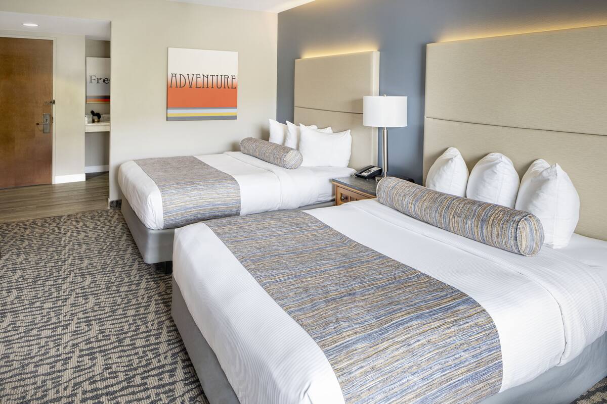 A hotel room with two beds, a nightstand, a lamp, and a wall-mounted artwork.