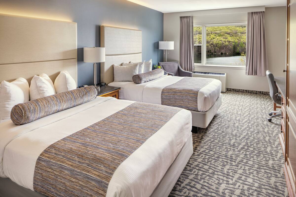 A hotel room with two beds, a large window with a view of greenery, a desk with a lamp, and a chair.