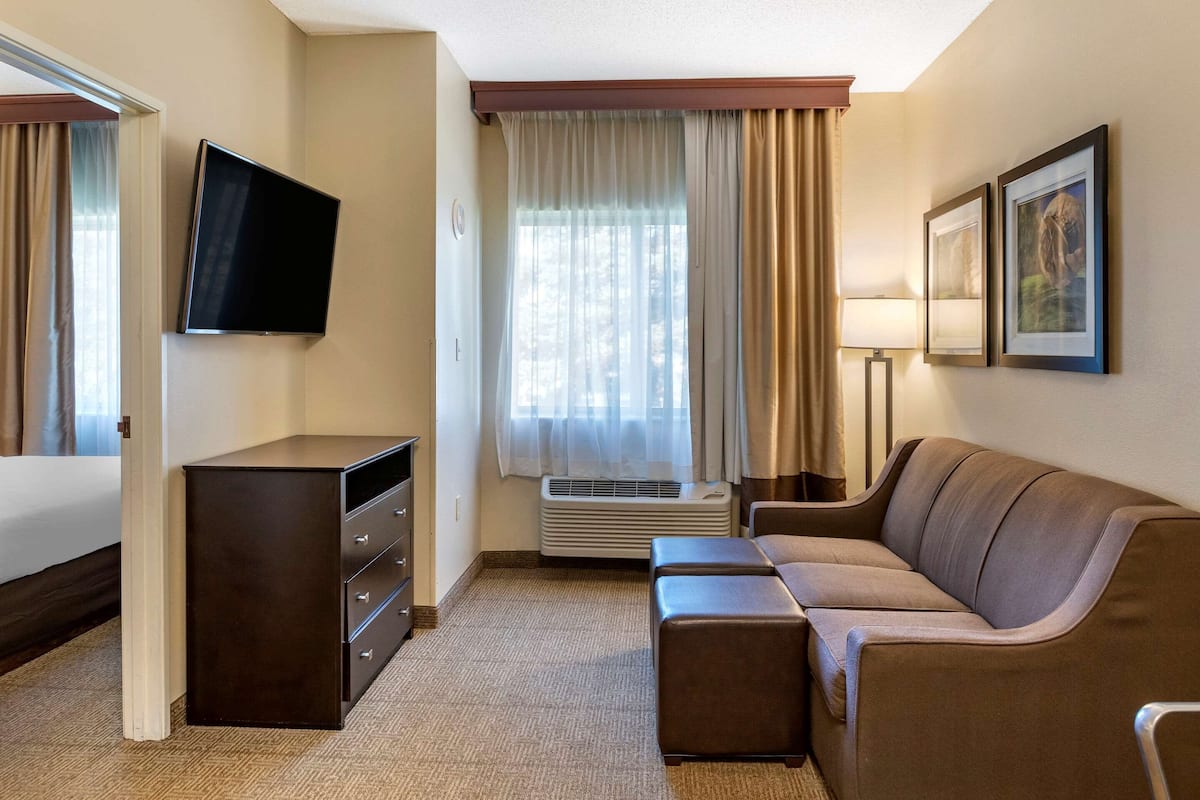 Ein Hotelzimmer mit Flachbildfernseher an der Wand, einer Couch, einem kleinen Tisch, einem Schminktisch und einem Fenster mit Vorhängen.