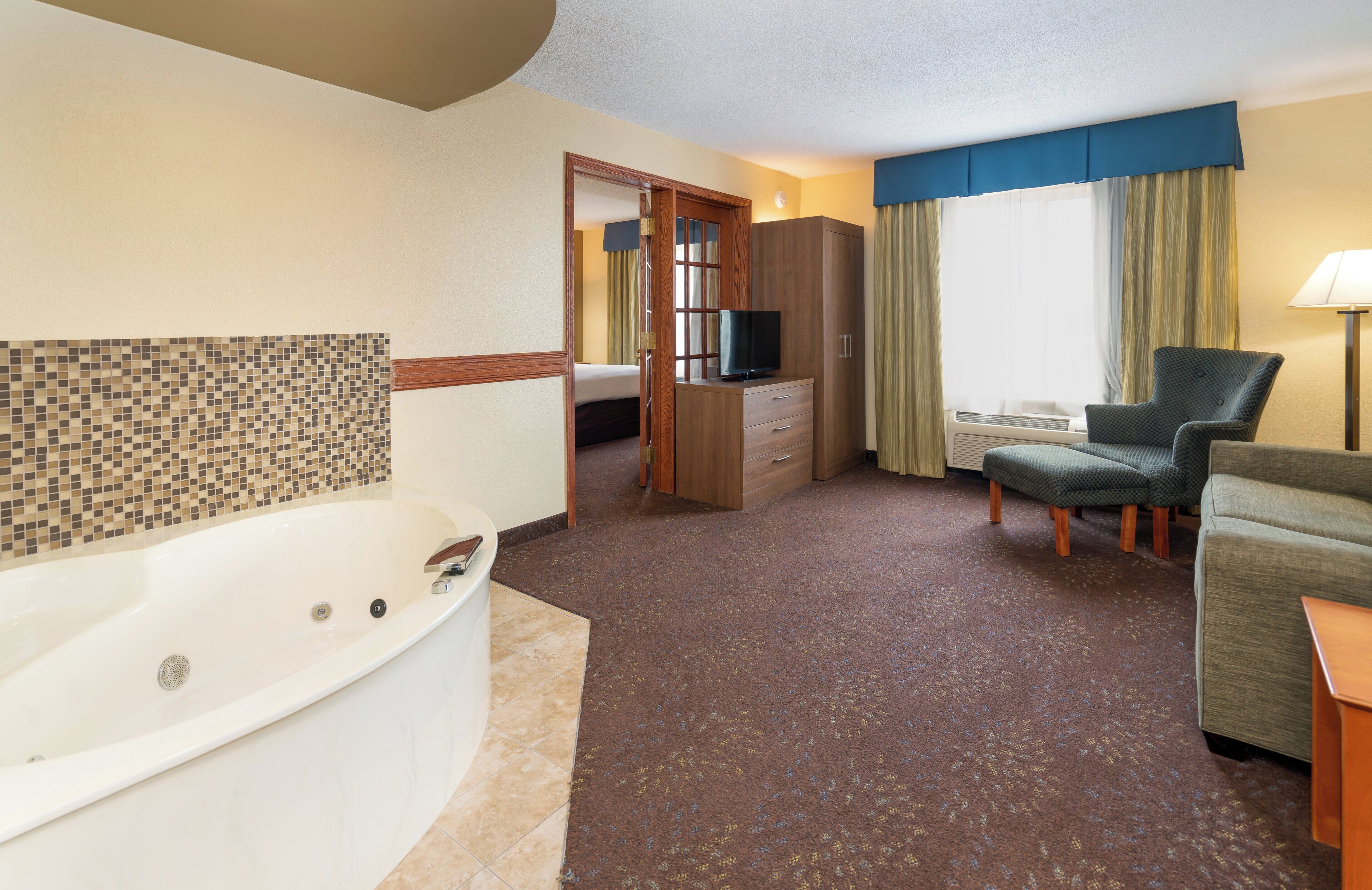 A hotel room with a bathtub, a television, a sofa, a chair, and a wardrobe.