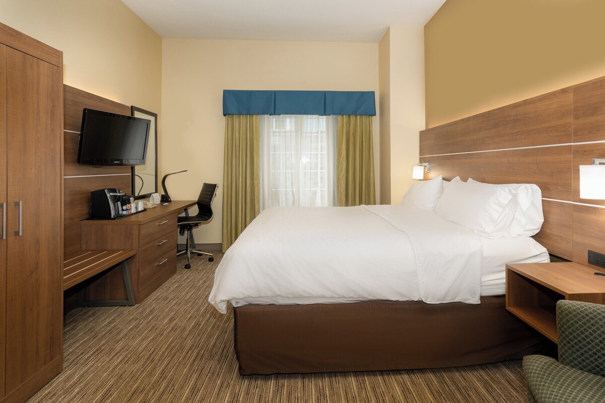A hotel room with a bed, a desk with a chair, a television, and a window with curtains.
