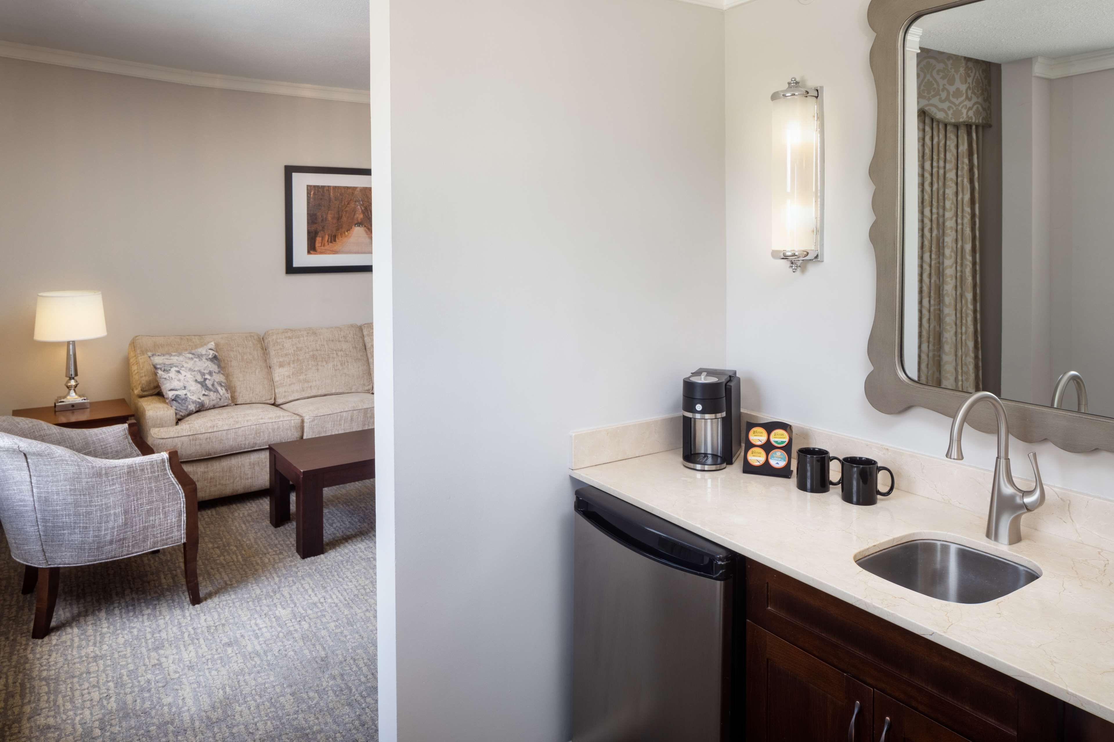 A hotel room with a sofa, chair, coffee maker, mugs, and a sink.