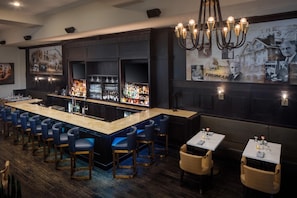 Eine Bar mit einer langen Theke, Barhocker und einer Wand mit gerahmten Bildern und einem Chandelier.