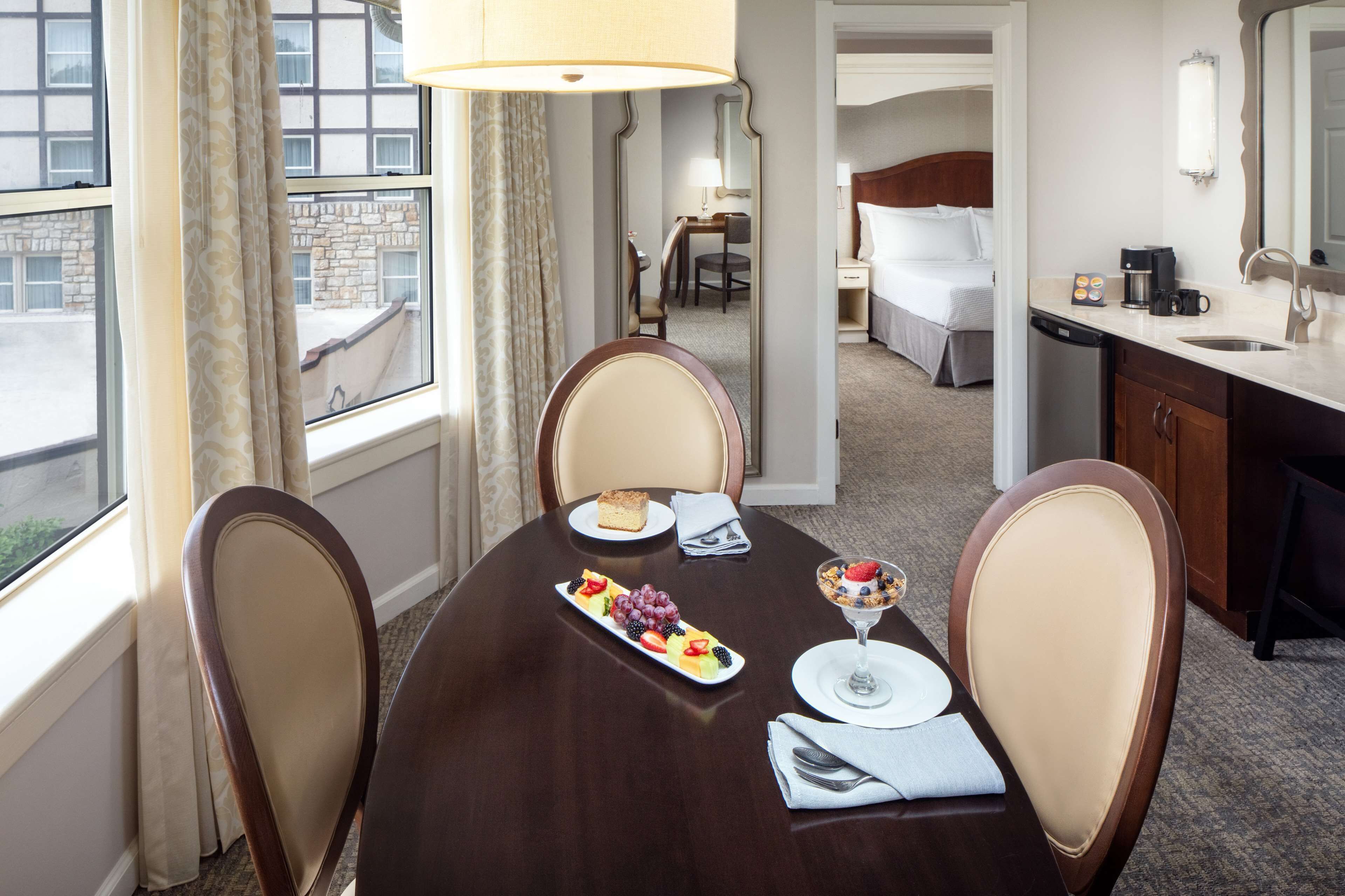 Habitación de hotel con mesa de comedor para dos, cama, baño y vistas al exterior.