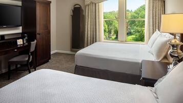A hotel room with two beds, a desk with a television, and a window with a view of greenery.