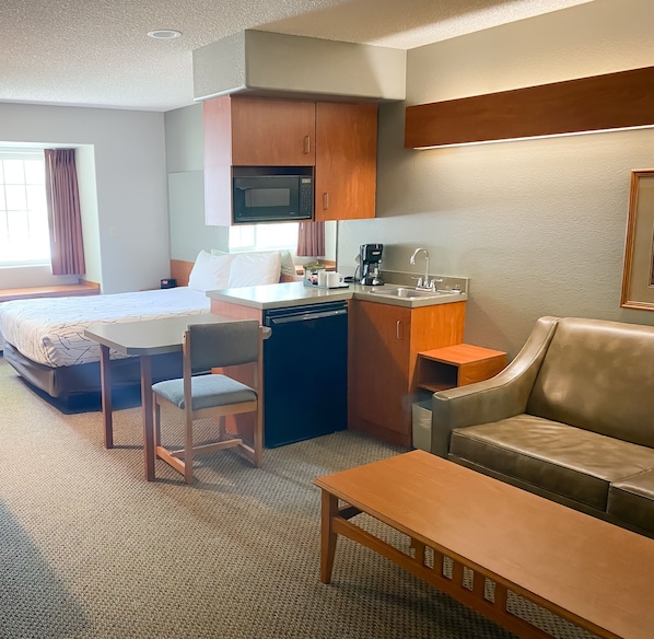 A compact hotel room with a bed, a small kitchenette, a dining area, and a seating area.