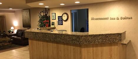 A hotel reception desk with a clock, a sign for the Monument Inn & Suites, and a potted plant.