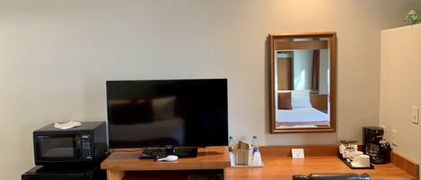 Una habitación de hotel con un escritorio de madera, televisión, microondas, cafetera y un cubo de basura.
