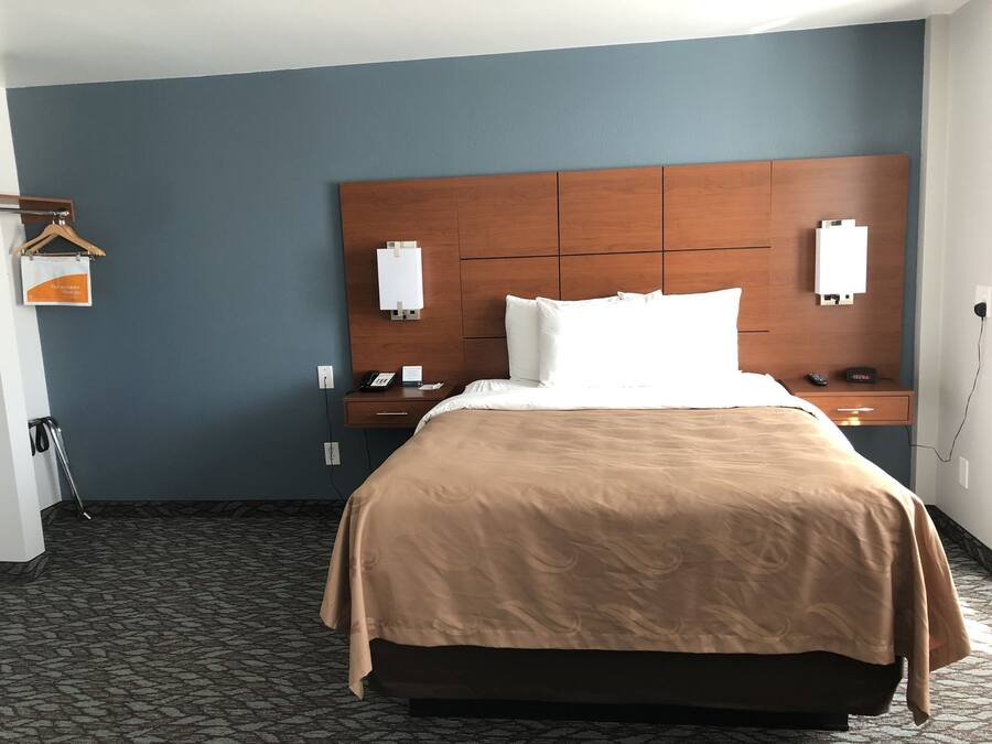 A hotel room with a bed, a wooden headboard, a nightstand with a phone and a remote, a wall-mounted light, and a carpeted floor.